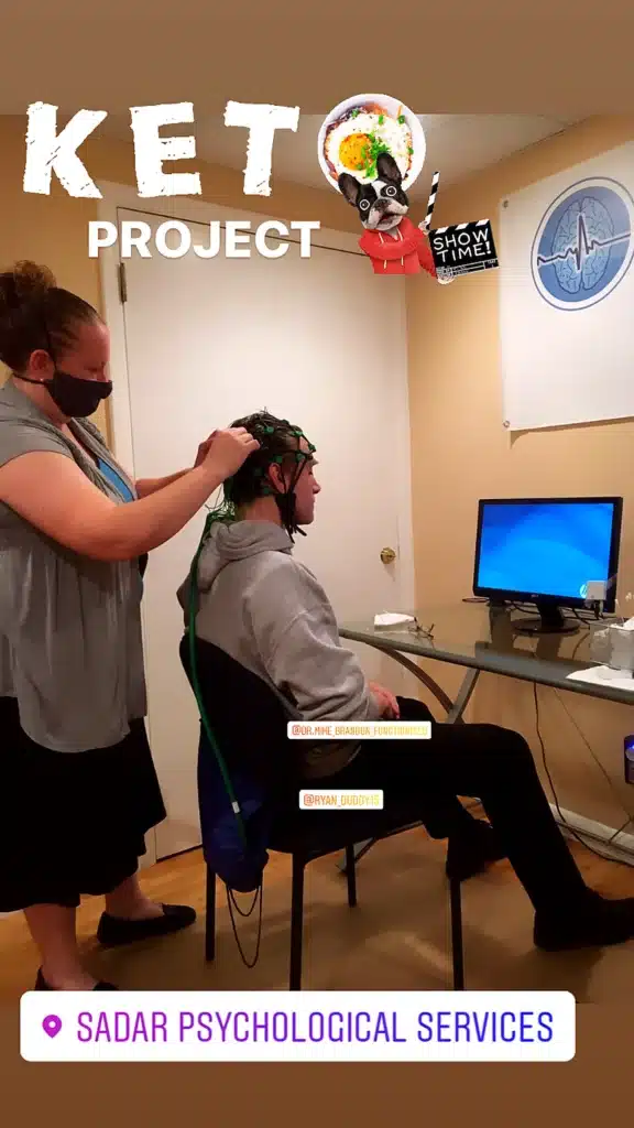 A person wearing a mask adjusts EEG electrodes on another person's head in a lab setting.