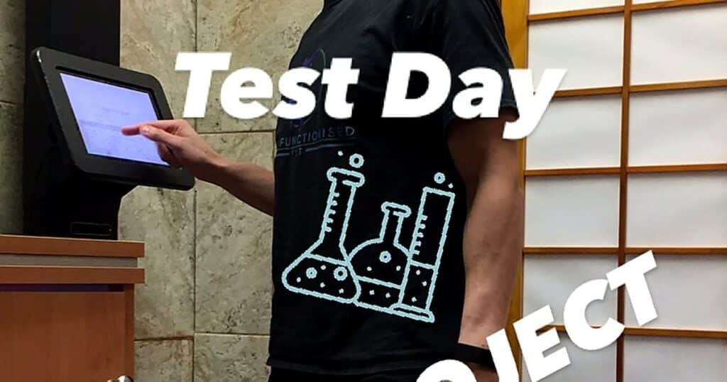 A person in a black t-shirt with a science-themed graphic stands next to a touchscreen kiosk, labeled 'Test Day.'