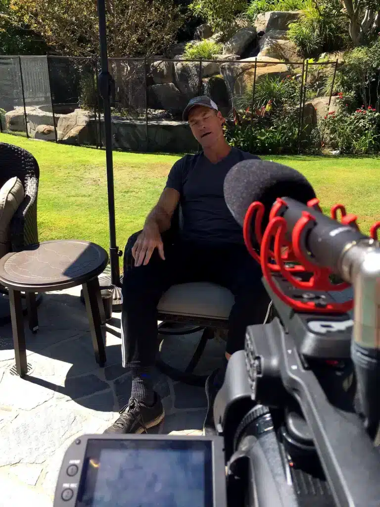 A man in a black shirt sits outdoors, speaking with a microphone aimed at him.