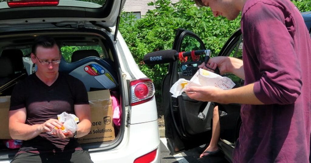 Two men sitting in a car's trunk area, one eating a burger while the other holds a microphone.