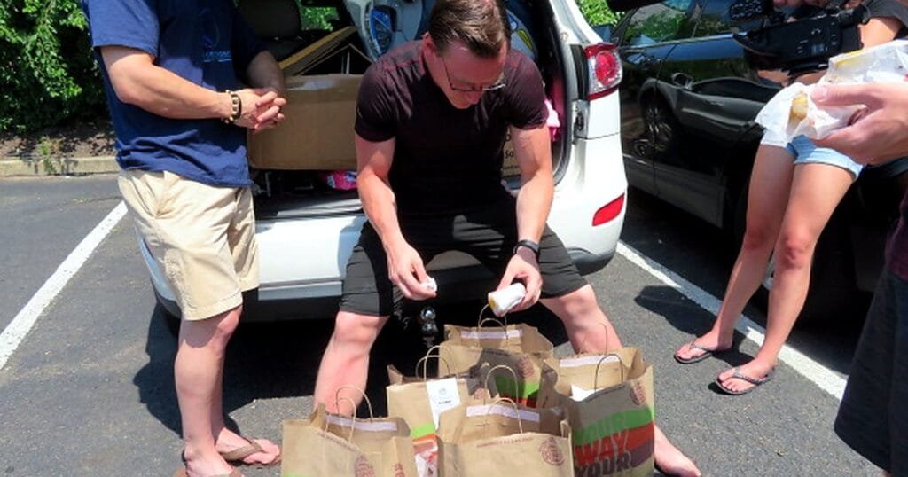 A man in sunglasses inspects a burger bag while sitting near a car filled with food bags.