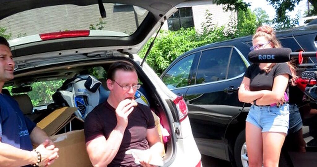 A man sits in a car trunk eating while another man stands nearby and a woman watches, with filming equipment visible.