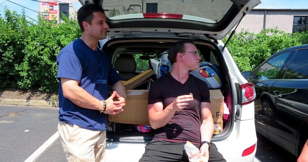 Two men stand by the open trunk of a car filled with boxes and supplies. One is eating, while the other is talking.