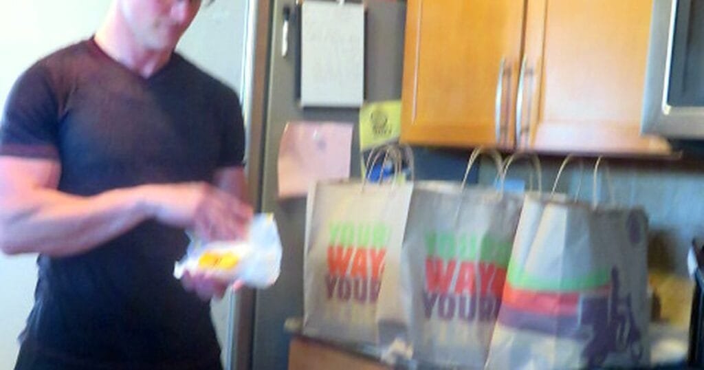 A fit man in a black shirt prepares food from takeout bags in a kitchen.