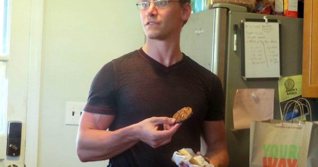 A fit man in a black shirt holds a burger while standing in a kitchen.