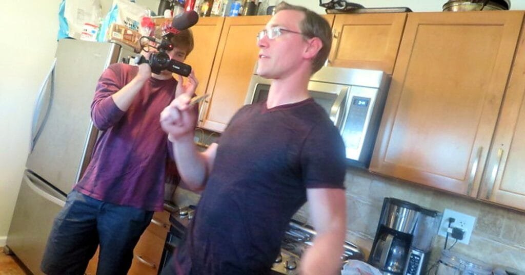 A man in a dark shirt speaks in a kitchen while another man films him with a camera.