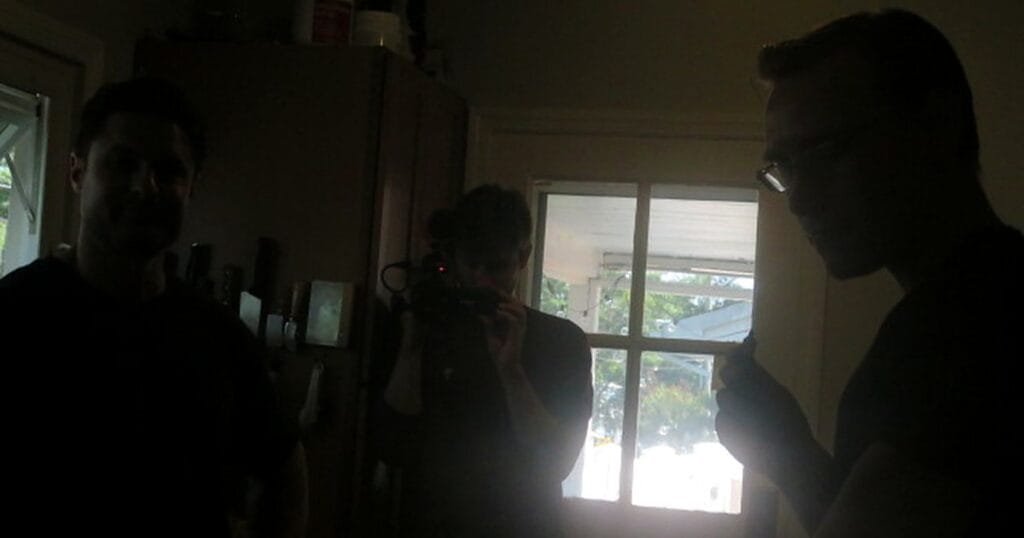 Two people in a dimly lit room, one holding a camera, the other holding an object, with a window in the background.