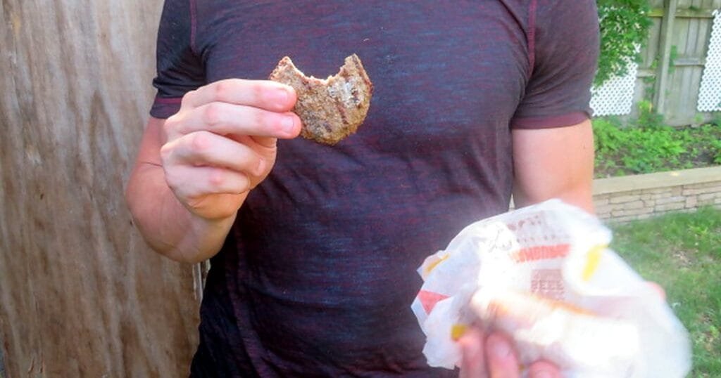 A person holds a half-eaten burger in one hand and a wrapper in the other, standing outdoors.