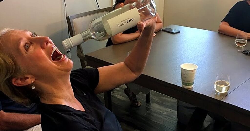 A woman tilts her head back, holding a wine bottle above her open mouth, while others sit around a table.