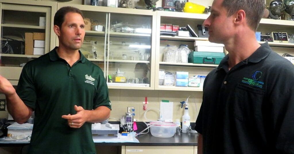 Two men in polo shirts talking in a lab filled with equipment and supplies.
