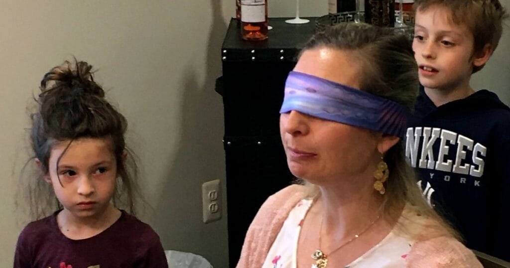 A woman with a blindfold stands in front of two children, one girl and one boy, observing her.