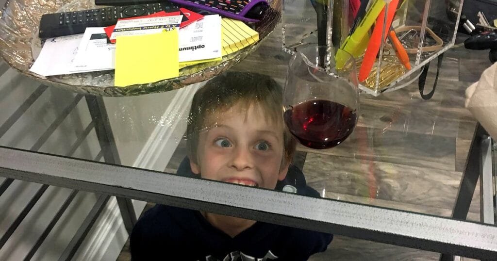 A child peeks under a glass table, making a funny face. A glass of red wine sits on the table.