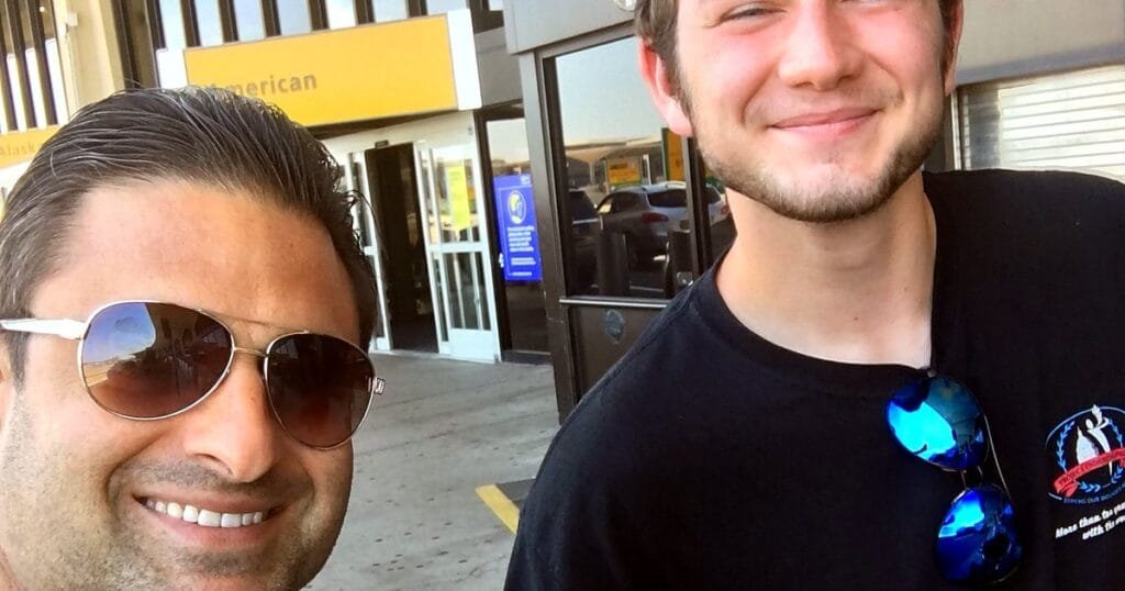 Two men smiling for a selfie outside a building with a yellow sign.