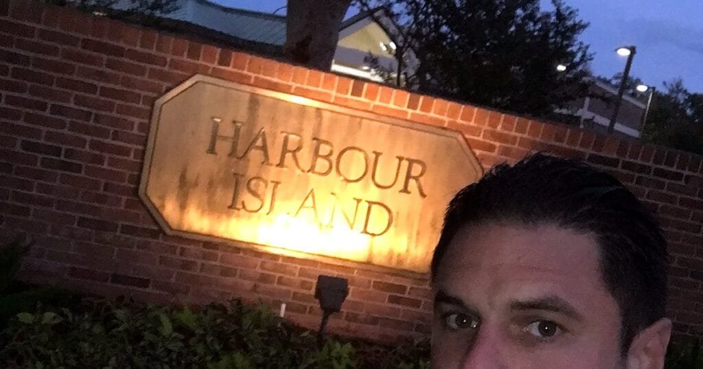 A man takes a selfie in front of a sign that reads 'Harbour Island' illuminated at night.