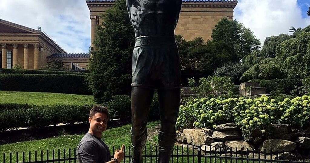 A person poses next to a bronze statue of a boxer with arms raised in victory, surrounded by greenery.