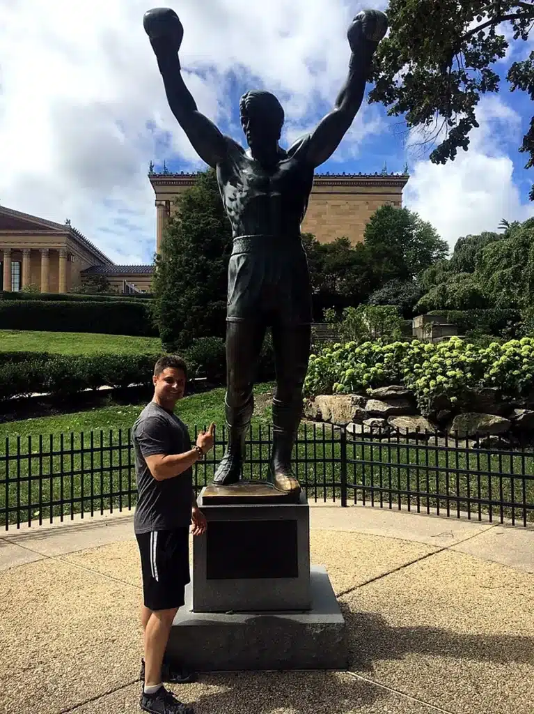A person poses next to a bronze statue of a boxer with arms raised in victory, surrounded by greenery.